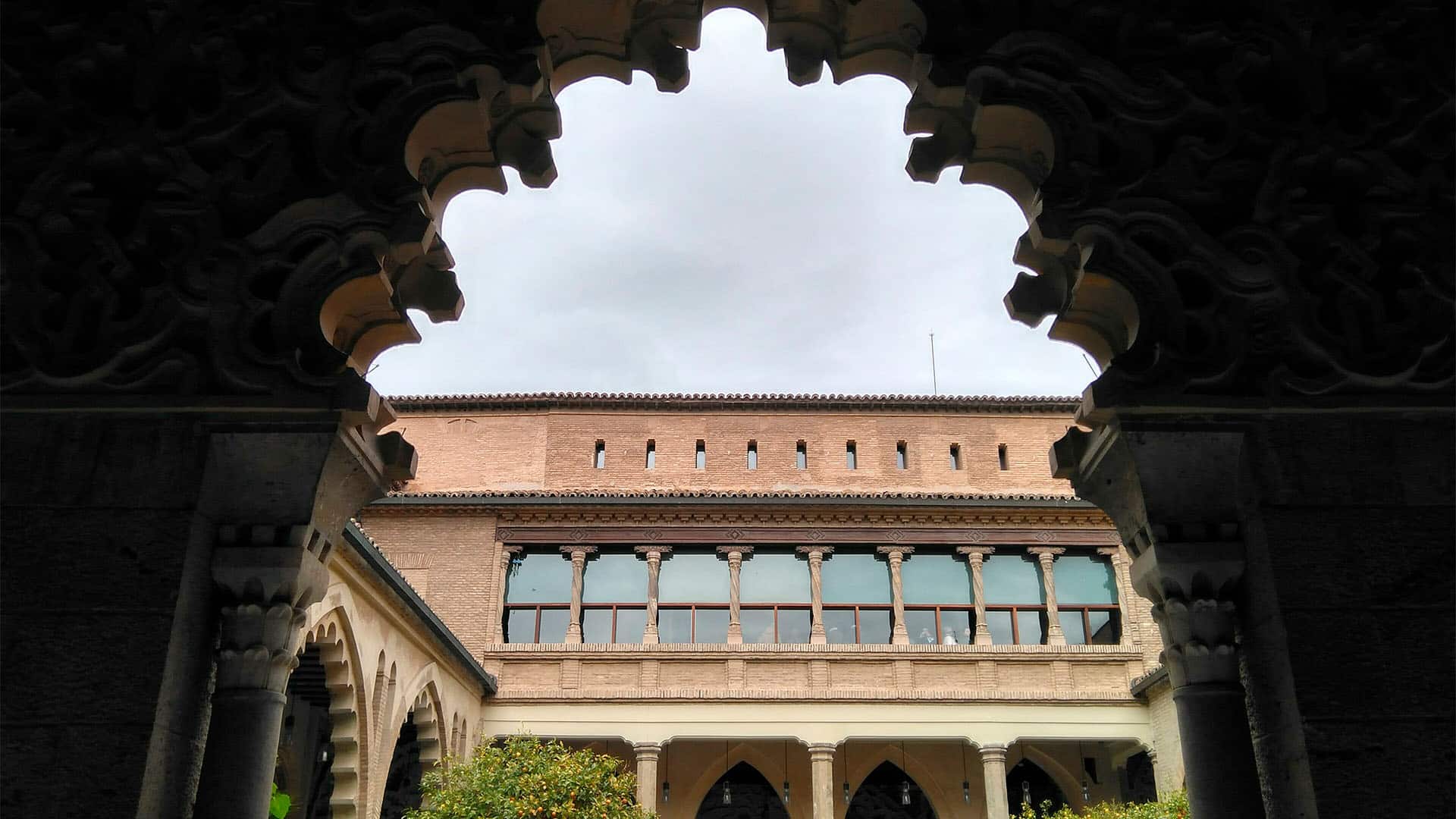Vista del Patio de Santa Isabel del Palacio de la Aljafería, en Zaragoza, enmarcado por un arco polilobulado en primer plano. Al fondo, una galería porticada con columnas, una fachada de ladrillo con un ventanal y vegetación en el patio bajo un cielo nublado.