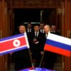 Vladimir Putin and Kim Jong-un enter a room, surrounded by several individuals. In the foreground, the flags of North Korea and Russia are seen in a blurred focus, symbolizing the diplomatic significance of the meeting.