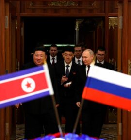 Vladimir Putin and Kim Jong-un enter a room, surrounded by several individuals. In the foreground, the flags of North Korea and Russia are seen in a blurred focus, symbolizing the diplomatic significance of the meeting.