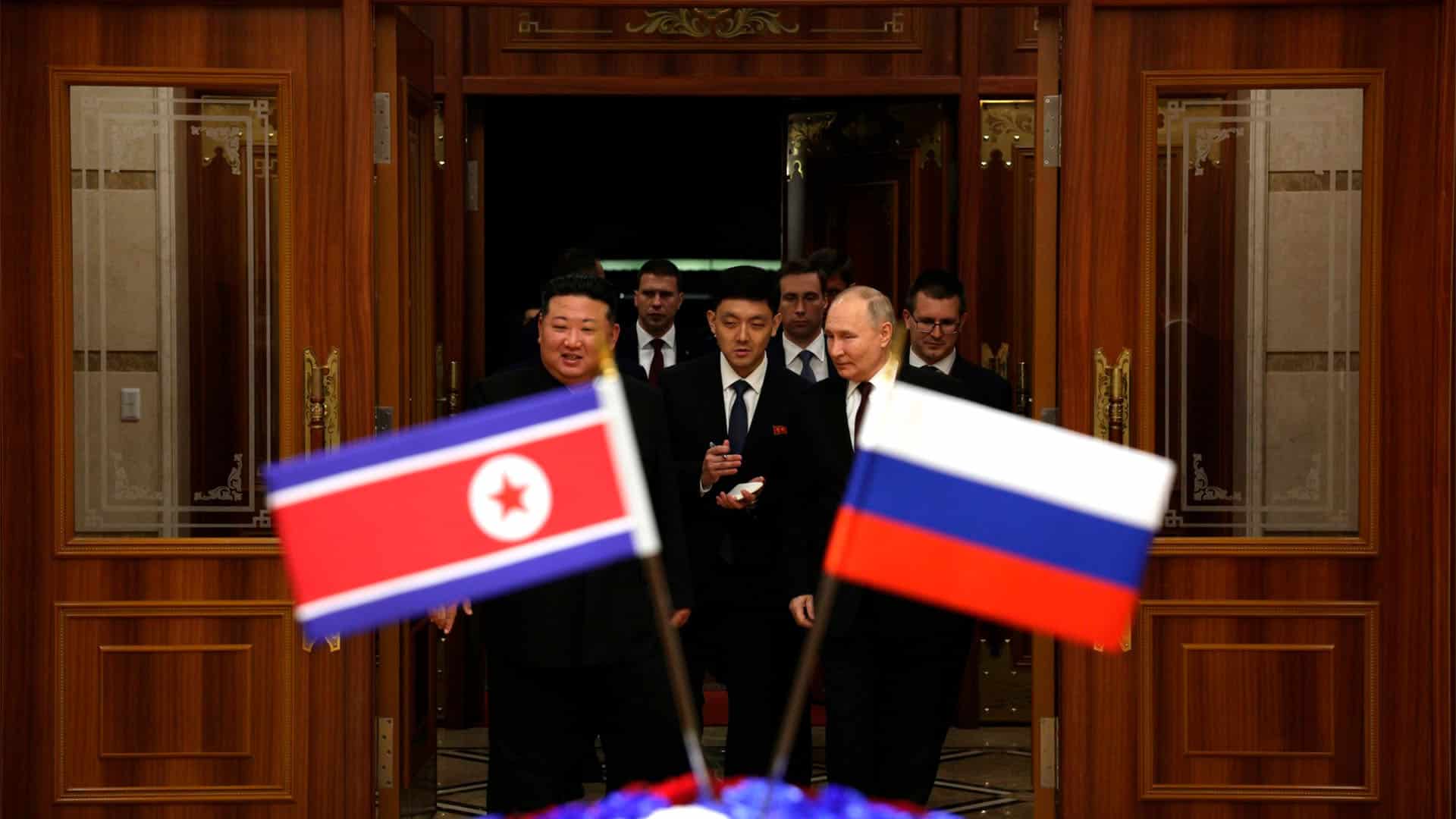 Vladimir Putin and Kim Jong-un enter a room, surrounded by several individuals. In the foreground, the flags of North Korea and Russia are seen in a blurred focus, symbolizing the diplomatic significance of the meeting.