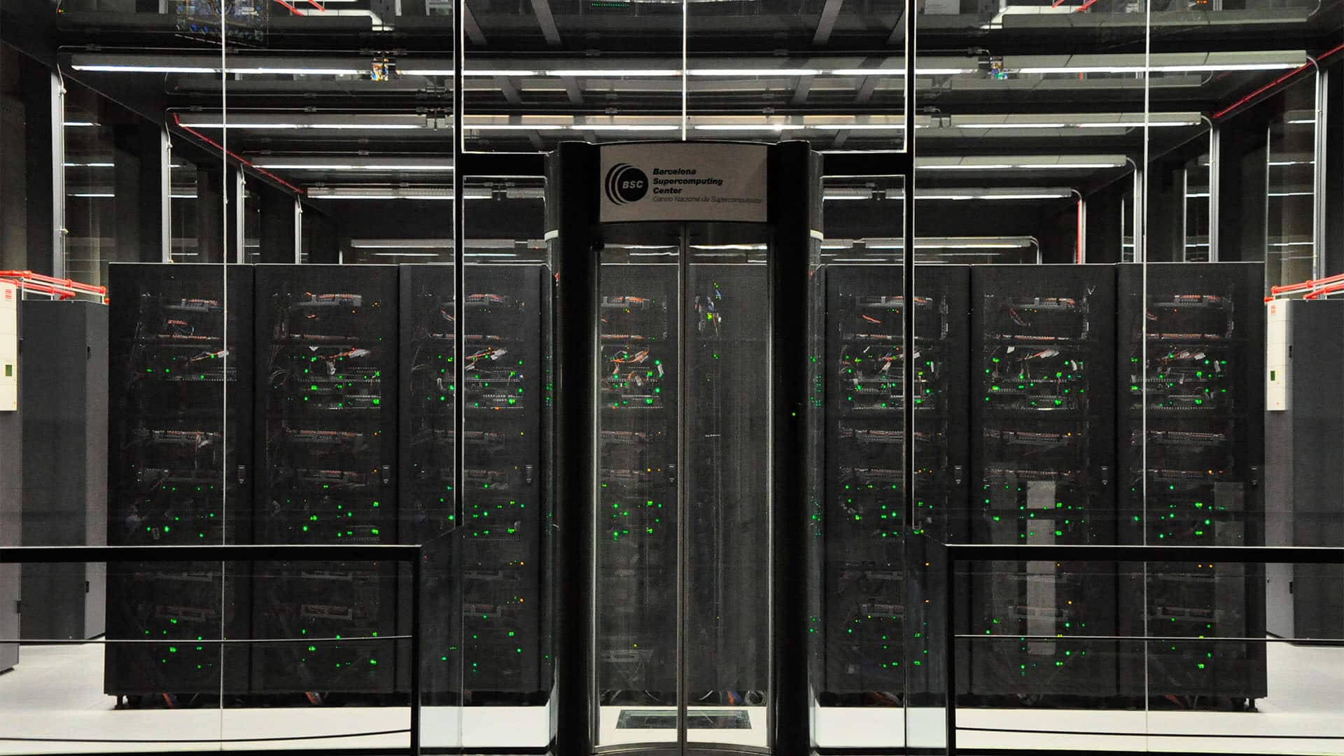 Front view of the MareNostrum supercomputer at the Barcelona Supercomputing Center: multiple rows of black server racks with blinking green lights, visible behind glass panels in a large, industrial-lit room.