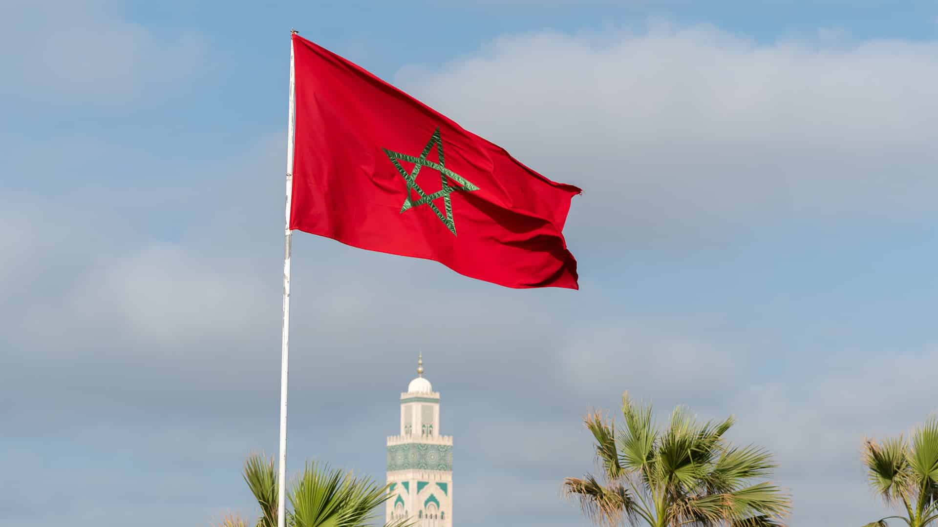 Bandera nacional de Marruecos ondeando frente al minarete de la mezquita Hassan II en Casablanca, Marruecos en un cielo claro y despejado.