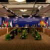Amplia sala de conferencias con techo modular de paneles claros, luminarias circulares y estructuras suspendidas de iluminación y sonido. El espacio tiene una alfombra oscura con patrones geométricos y varias plantas ornamentales verdes con flores rojas distribuidas en el centro. A ambos lados, representantes diplomáticos están sentados alrededor de una mesa en forma de U durante una reunión oficial de UNASUR en Perú, con banderas de países sudamericanos y pantallas institucionales al fondo.