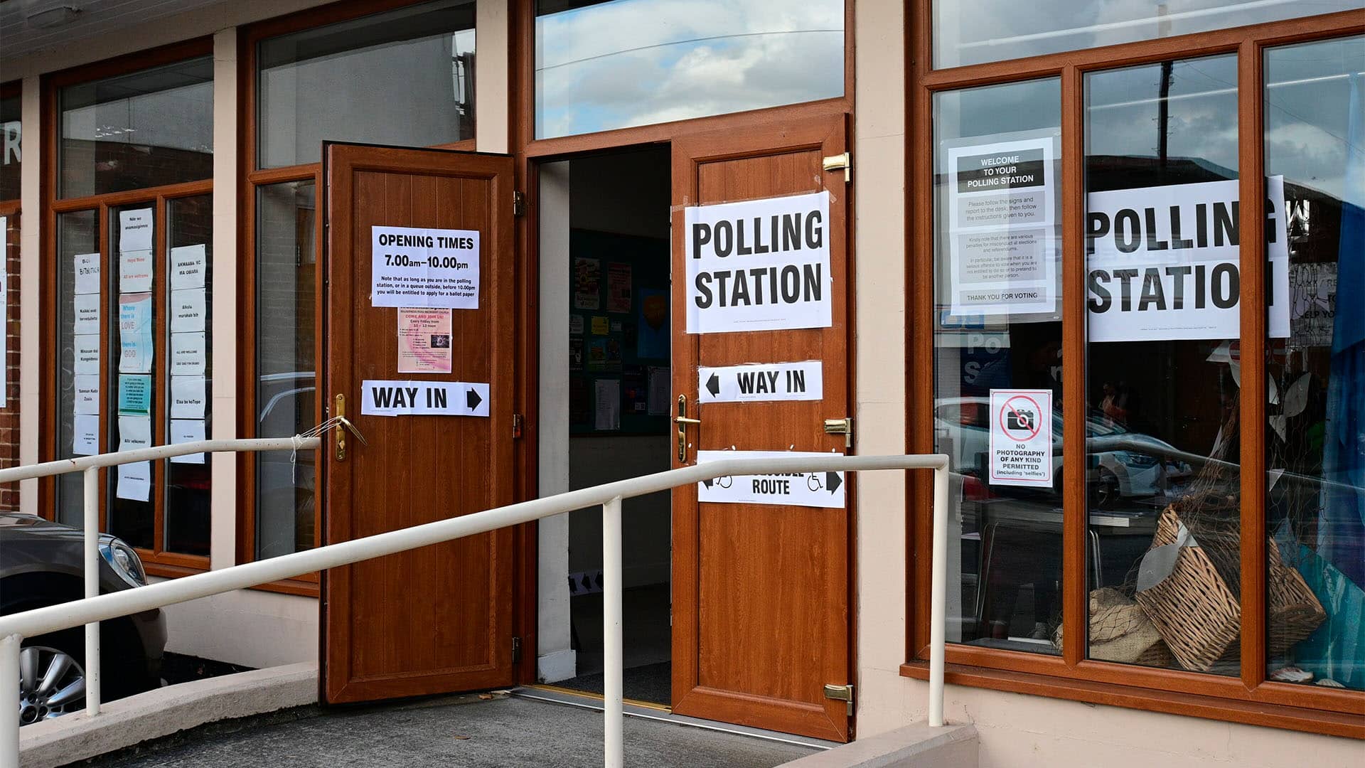 Entrance to a polling station in the Kingston upon Hull East constituency during the 2024 United Kingdom general election. Open wooden doors display large “Polling Station” and “Way In” signs, while an accessible ramp with white handrails leads to the entrance. Notices and election information are posted on the windows and doors, and reflections of the street and parked cars can be seen in the glass.