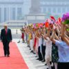 El presidente chino, Xi Jinping, y el presidente de EEUU, Donald Trump, caminan por una alfombra roja durante una ceremonia oficial en Pekín mientras un grupo de jóvenes agita banderas de China y Estados Unidos y sostiene ramos de flores. Al fondo se distingue el Gran Palacio del Pueblo y un monumento en la plaza de Tiananmén.