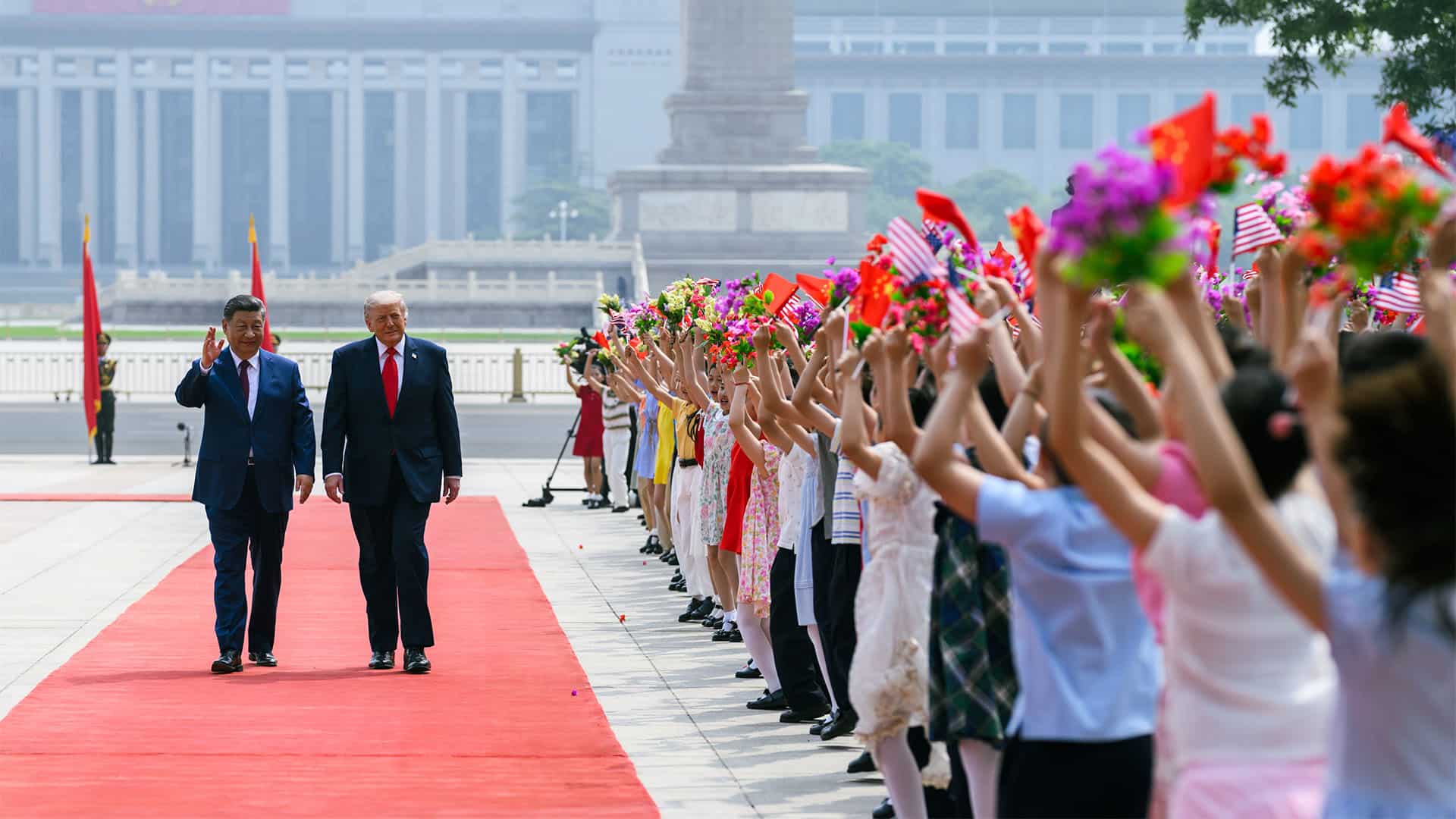 El presidente chino, Xi Jinping, y el presidente de EEUU, Donald Trump, caminan por una alfombra roja durante una ceremonia oficial en Pekín mientras un grupo de jóvenes agita banderas de China y Estados Unidos y sostiene ramos de flores. Al fondo se distingue el Gran Palacio del Pueblo y un monumento en la plaza de Tiananmén.