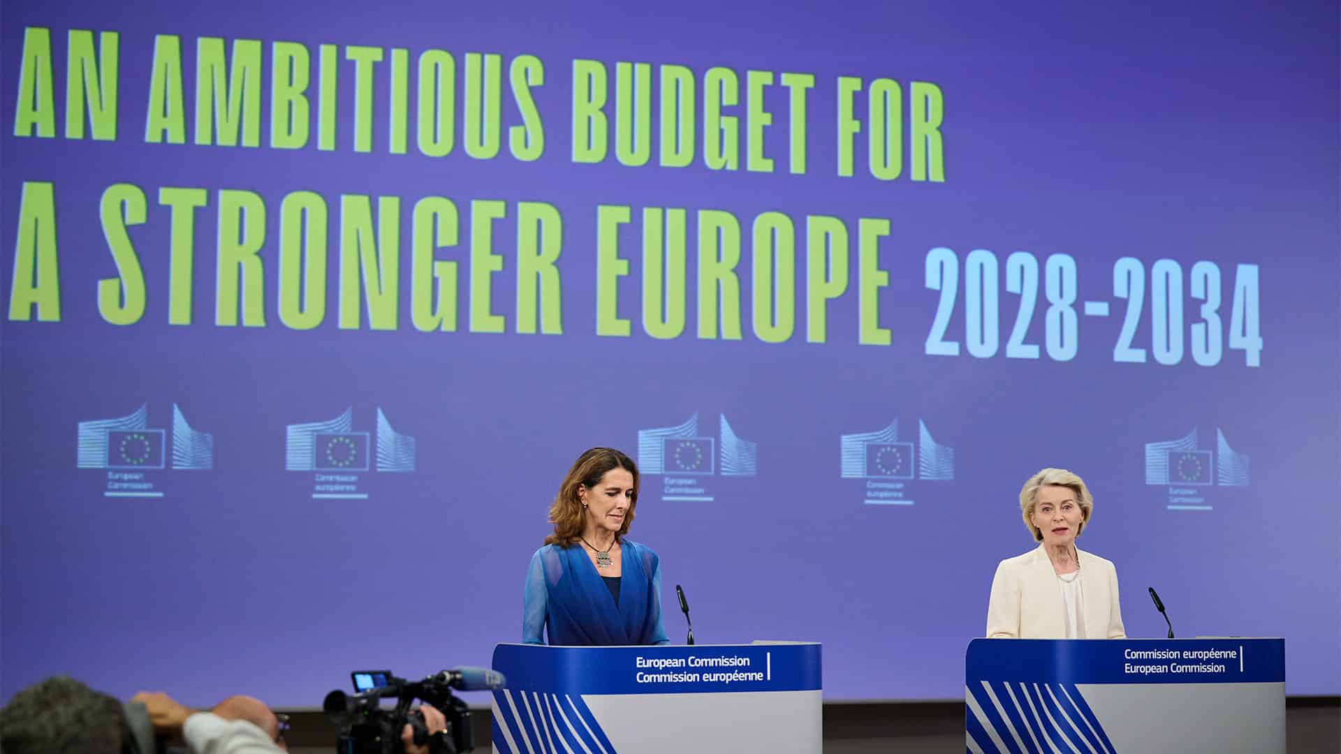 Paula Pinho, portavoz principal de la Comisión Europea (a la izquierda) y Ursula von der Leyen, presidenta de la Comisión Europea (a la derecha) hablando desde los atriles ante la prensa durante el resumen de la reunión semanal de la Comisión sobre el próximo presupuesto de la UE. A la izquierda se asoma una cámara de televisión y al fondo la pantalla azul proyecta el logo de la Comisión en fila y las palabras en inglés “An ambitious budget for a stronger Europe 2028-2034” (un presupuesto ambicioso para una Europa más fuerte 2028-2034).