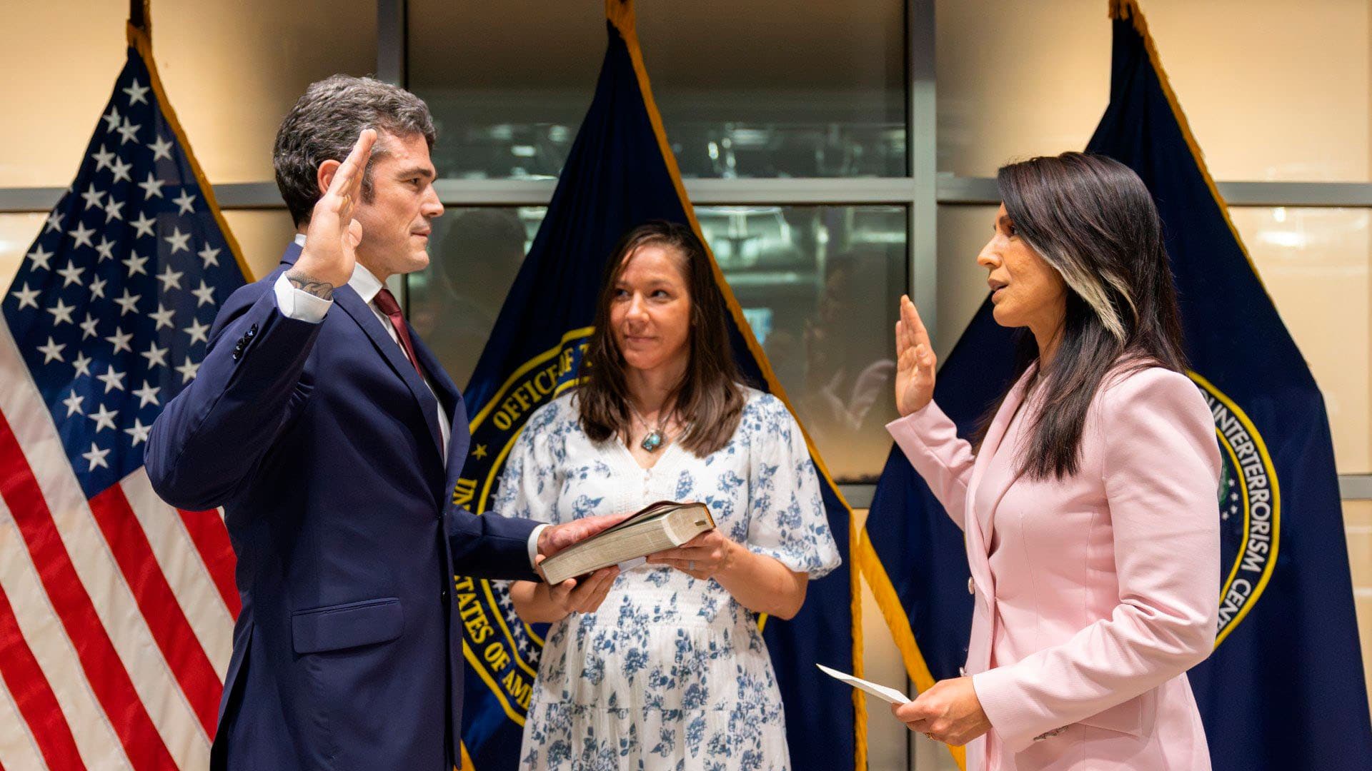 Joe Kent presta juramento como director del National Counterterrorism Center (NCTC) de Estados Unidos ante la directora de Inteligencia Nacional, Tulsi Gabbard, el 31 de julio de 2025. Kent aparece a la izquierda con la mano derecha levantada mientras sostiene un libro con la izquierda. Frente a él, Gabbard también levanta la mano derecha y sostiene un documento. Entre ambos, una mujer actúa como testigo de la ceremonia. Al fondo se ven tres banderas: la de Estados Unidos, la de la Oficina de Inteligencia Nacional y la del Centro Nacional Antiterrorista.