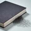 Closed black hardcover book on a light surface, with scattered letters emerging from the edge of the pages, creating the visual effect of words disintegrating.