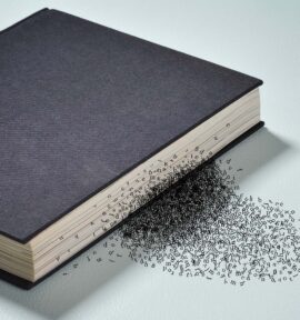 Closed black hardcover book on a light surface, with scattered letters emerging from the edge of the pages, creating the visual effect of words disintegrating.