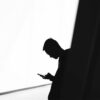 Silueta de un joven mirando la pantalla de un teléfono móvil en un espacio interior de diseño minimalista. La imagen en blanco y negro muestra un fuerte contraste entre las paredes iluminadas y las sombras.