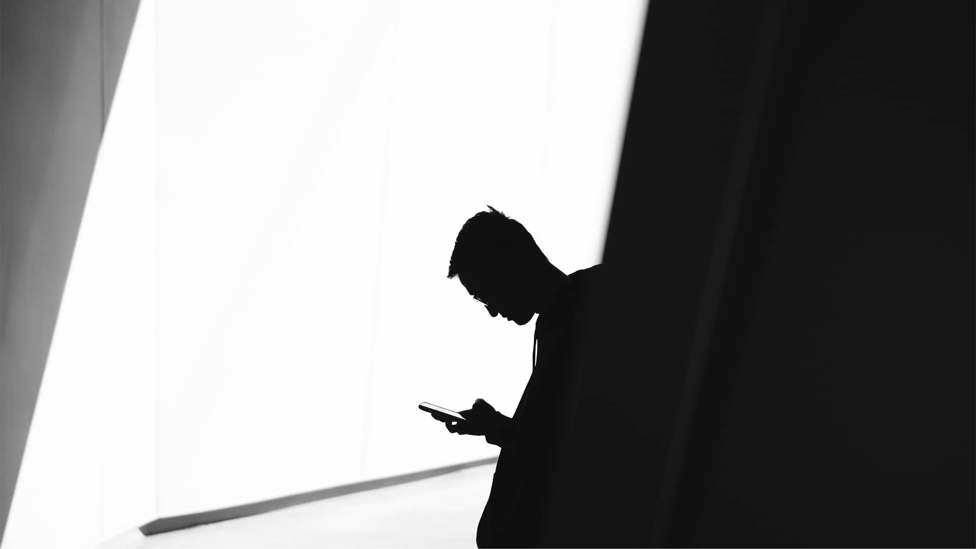 Silueta de un joven mirando la pantalla de un teléfono móvil en un espacio interior de diseño minimalista. La imagen en blanco y negro muestra un fuerte contraste entre las paredes iluminadas y las sombras.