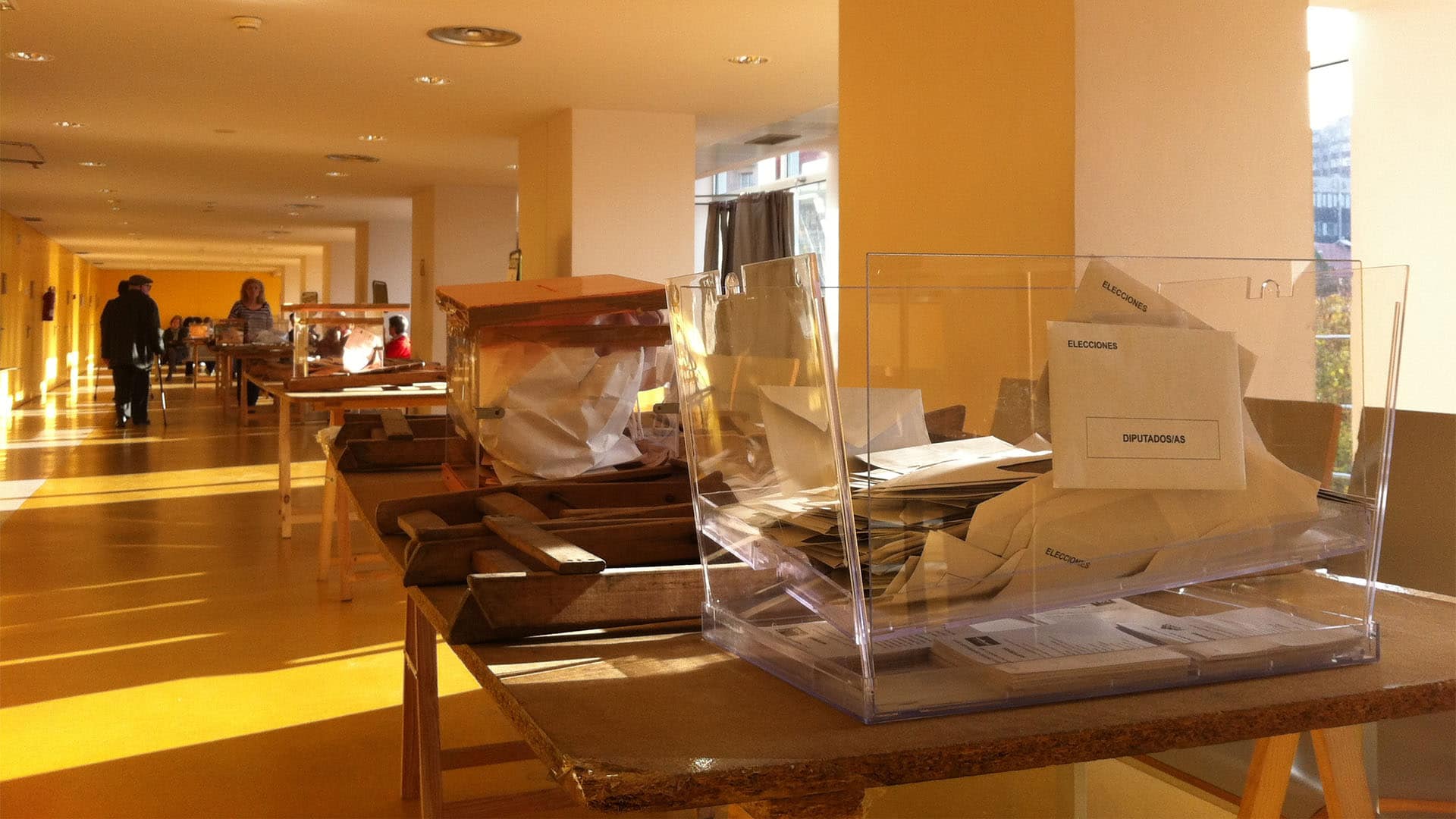 Transparent ballot boxes containing voting envelopes and ballots at a polling station in Spain. Voting tables and several people participating in the election process can be seen in the background.