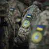 Primer plano de varios soldados de las Fuerzas Armadas de Ucrania con uniformes de camuflaje durante un acto oficial. En las mangas se aprecian parches con la bandera ucraniana y distintivos militares, mientras participan en un acto institucional encabezado por el presidente Volodímir Zelenski con motivo del Día de las Fuerzas Armadas de Ucrania.
