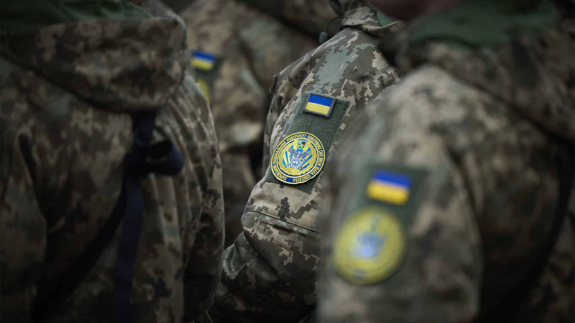 Primer plano de varios soldados de las Fuerzas Armadas de Ucrania con uniformes de camuflaje durante un acto oficial. En las mangas se aprecian parches con la bandera ucraniana y distintivos militares, mientras participan en un acto institucional encabezado por el presidente Volodímir Zelenski con motivo del Día de las Fuerzas Armadas de Ucrania.