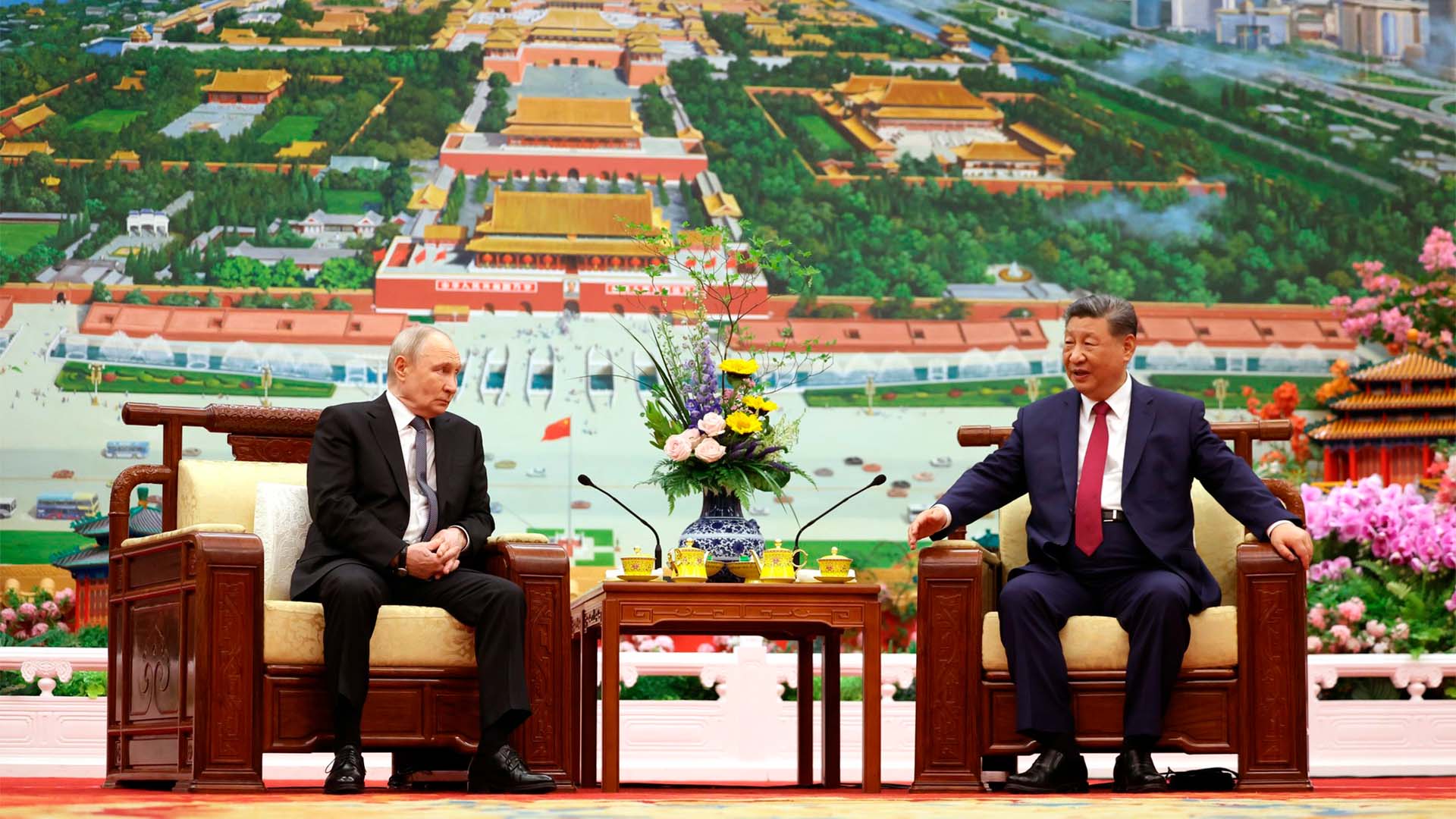 El presidente de Rusia, Vladímir Putin, y el presidente de China, Xi Jinping, mantienen una reunión oficial sentados frente a frente en un salón de Pekín. Entre ambos hay una mesa con un arreglo floral y servicio de té, mientras al fondo se observa una gran representación panorámica de la Ciudad Prohibida.