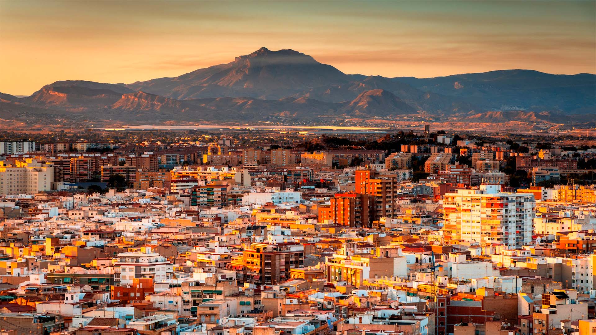 Panorámica de la ciudad de Alicante iluminada por la luz del atardecer, con edificios residenciales en primer plano y el monte Benacantil elevándose al fondo bajo un cielo despejado.