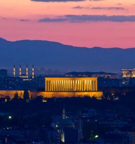 Vista panorámica de Ankara al atardecer con el mausoleo de Atatürk (Anıtkabir) iluminado en el centro de la ciudad. Al fondo se distinguen colinas, mezquitas y edificios bajo un cielo de tonos rosados y azulados.