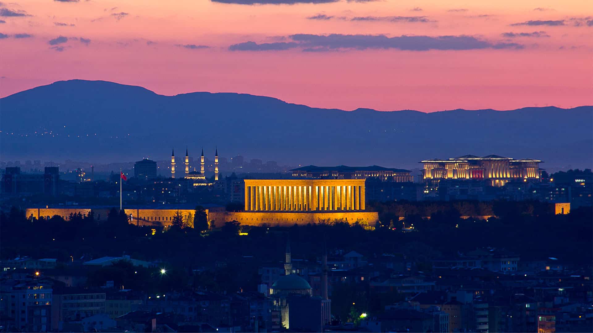 Panoramic view of Ankara at sunset featuring the illuminated mausoleum of Atatürk (Anıtkabir) at the city’s center. Hills, mosques, and urban buildings can be seen in the background beneath a pink and blue evening sky.