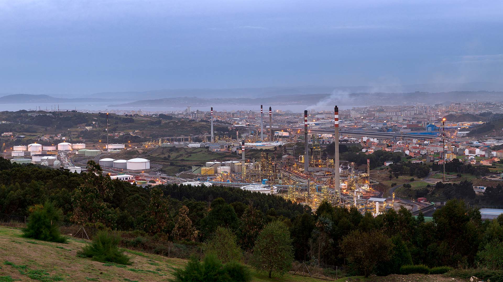 Panorámica de la refinería de petróleo de A Coruña al atardecer, con instalaciones industriales, depósitos de combustible y el entorno urbano y costero al fondo.