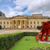 Vista frontal de la Casa de Nariño, sede de la Presidencia de Colombia en Bogotá, con banderas nacionales colgadas en su fachada neoclásica. En primer plano destaca la escultura roja Vigilantes, del artista Edgar Negret, rodeada de jardines y zonas verdes bajo un cielo parcialmente nublado.