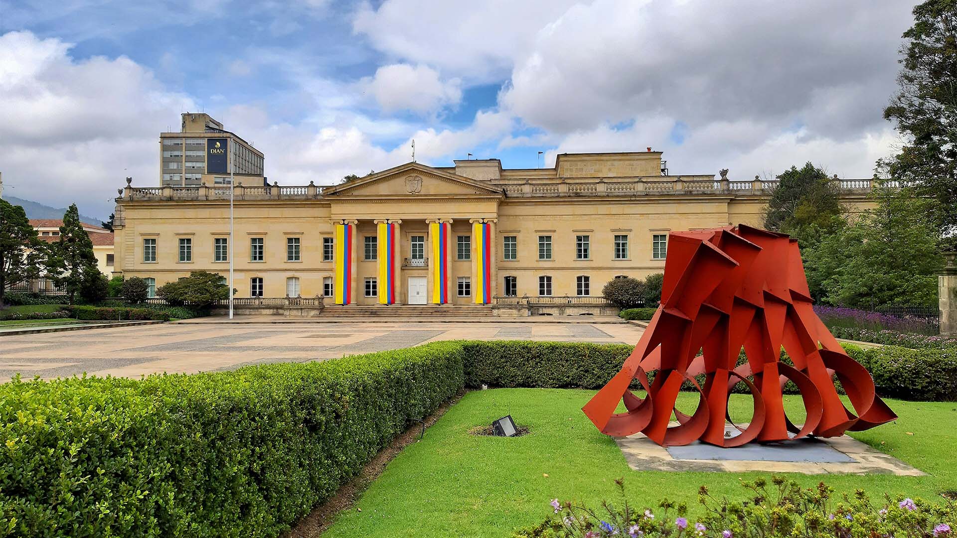Vista frontal de la Casa de Nariño, sede de la Presidencia de Colombia en Bogotá, con banderas nacionales colgadas en su fachada neoclásica. En primer plano destaca la escultura roja Vigilantes, del artista Edgar Negret, rodeada de jardines y zonas verdes bajo un cielo parcialmente nublado.