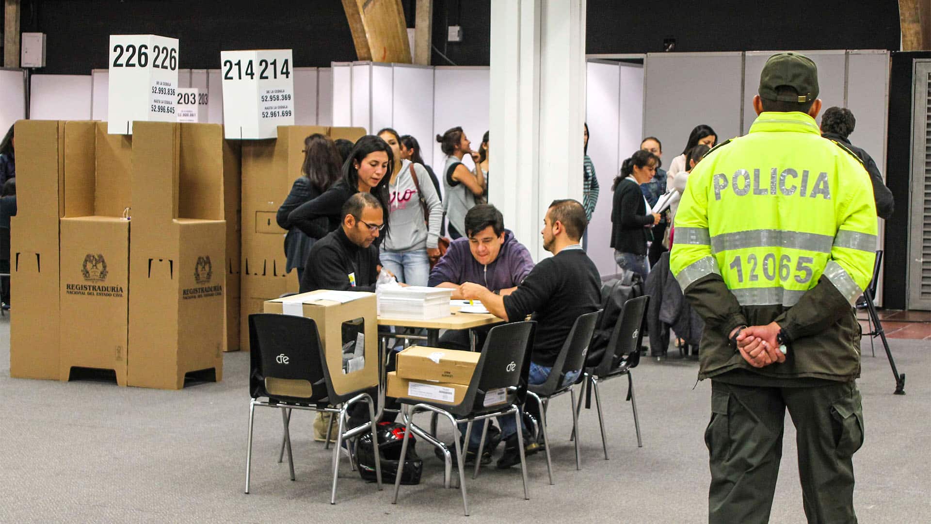 Interior de un centro de votación instalado en Corferias, Bogotá, con cabinas electorales, mesas de trabajo y personal realizando tareas de organización y recuento de votos, mientras un agente de la Policía Nacional supervisa la seguridad de la jornada electoral.