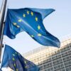 Varias banderas de la Unión Europea ondean a media asta frente al edificio Berlaymont, sede de la Comisión Europea en Bruselas, con la fachada acristalada del complejo institucional al fondo.