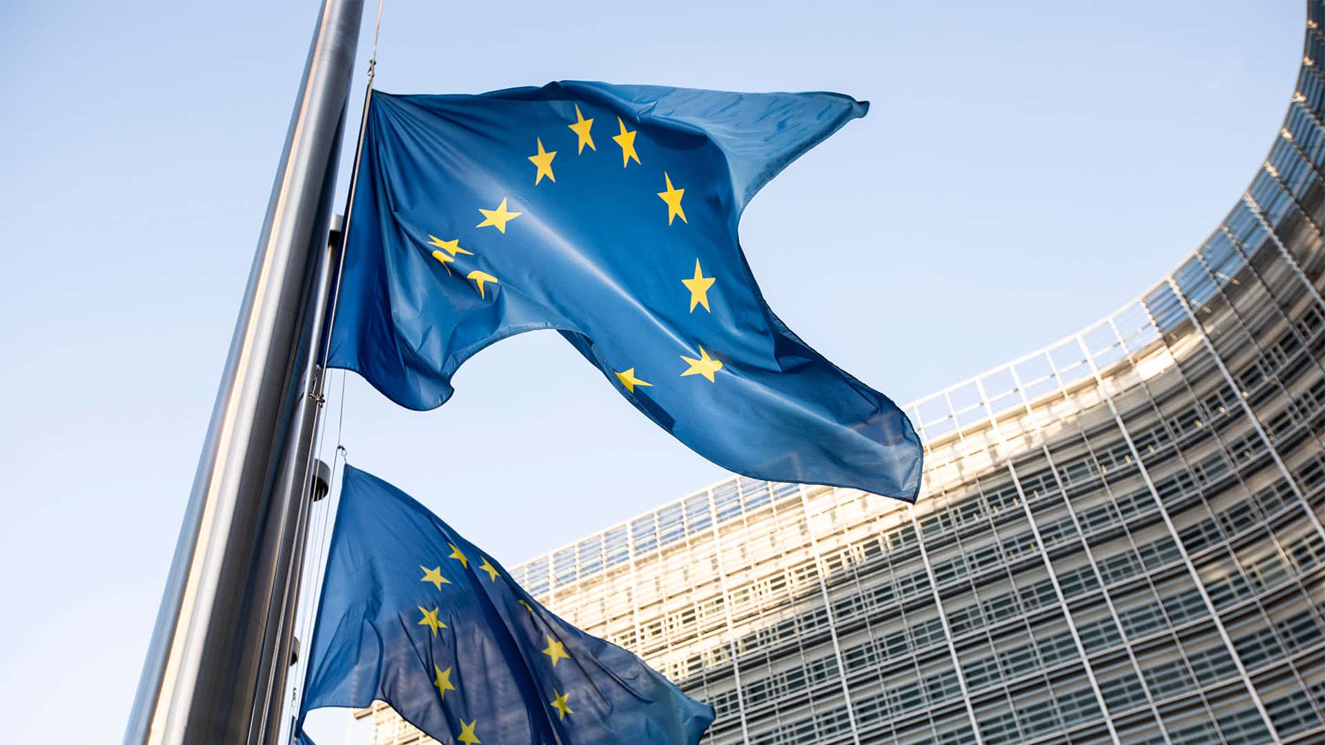 Varias banderas de la Unión Europea ondean a media asta frente al edificio Berlaymont, sede de la Comisión Europea en Bruselas, con la fachada acristalada del complejo institucional al fondo.