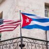 Banderas de Cuba y Estados Unidos ondeando juntas en la fachada de un edificio histórico de La Habana, con balcones de hierro forjado y ventanas de estilo colonial al fondo.
