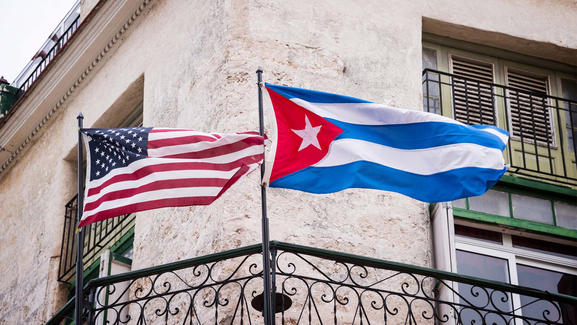 Banderas de Cuba y Estados Unidos ondeando juntas en la fachada de un edificio histórico de La Habana, con balcones de hierro forjado y ventanas de estilo colonial al fondo.