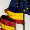 Banderas de España, Alemania y la Unión Europea ondean juntas en mástiles al aire libre, sobre un fondo blanquecino.