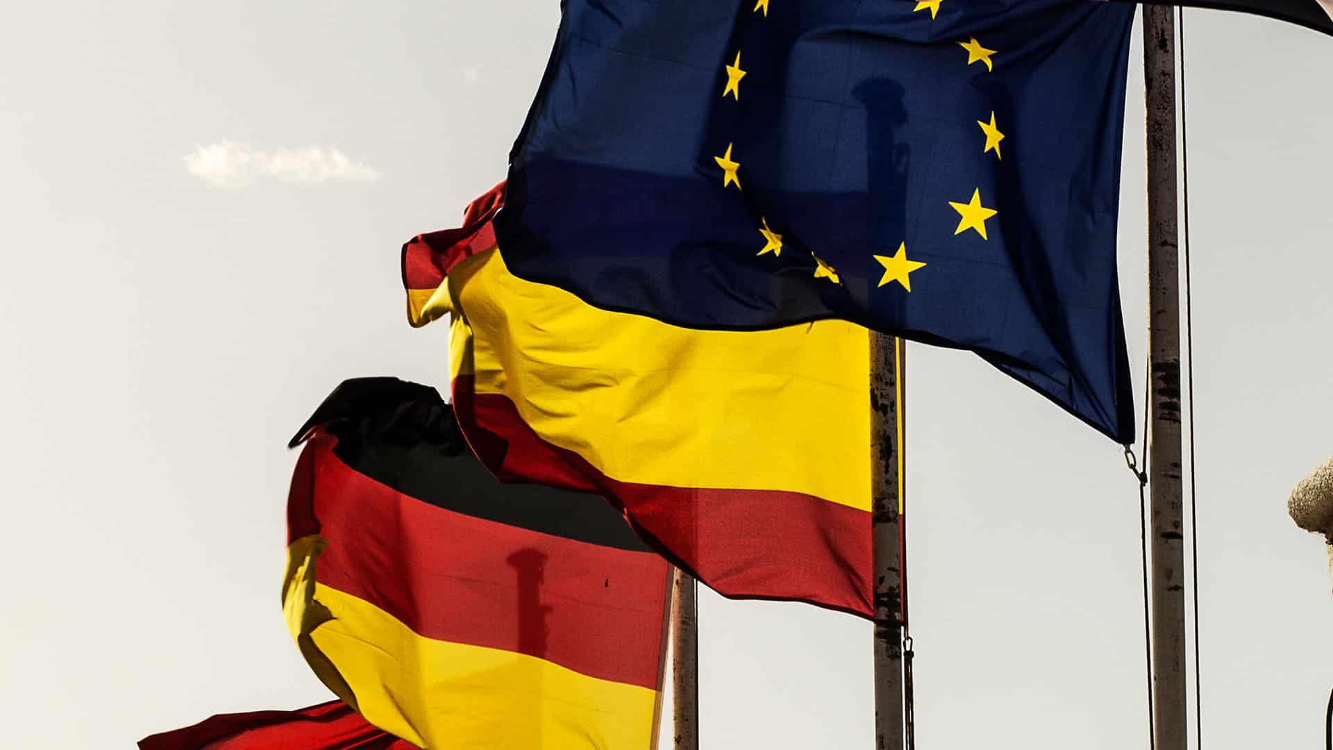 Banderas de España, Alemania y la Unión Europea ondean juntas en mástiles al aire libre, sobre un fondo blanquecino.