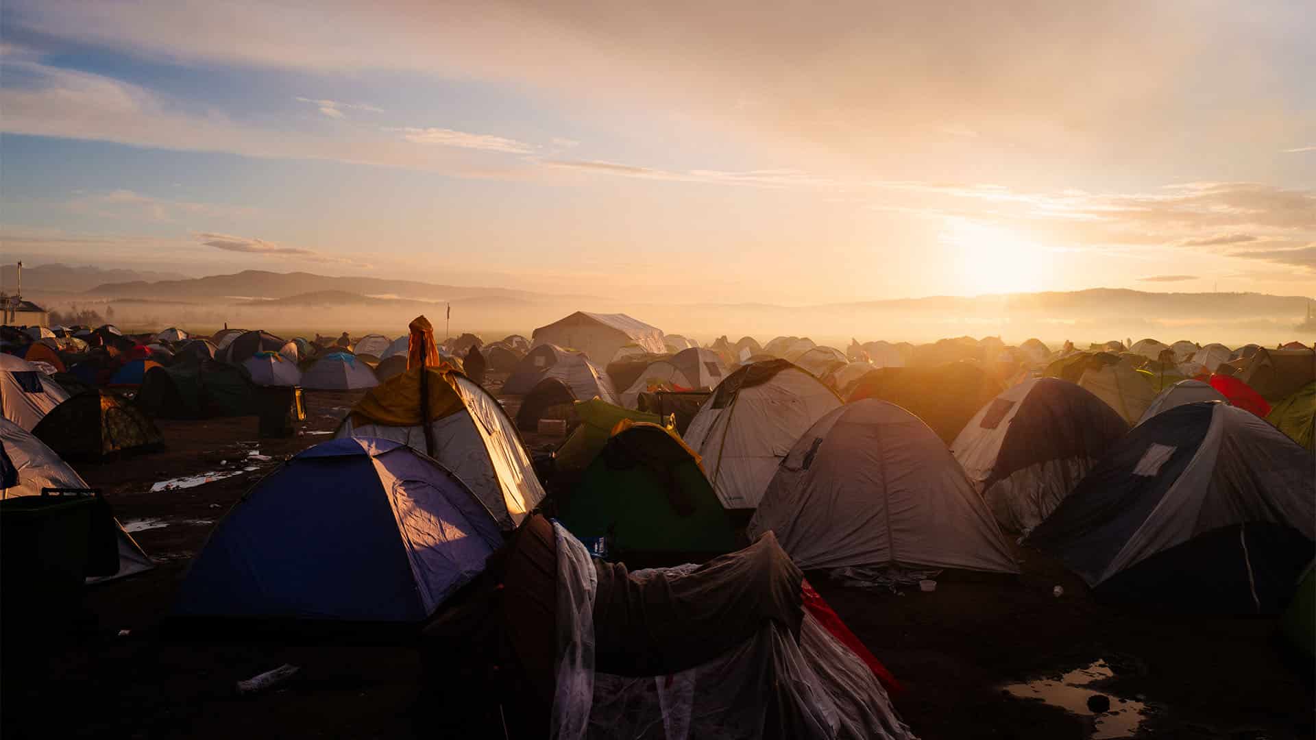 Campo de refugiados lleno de tiendas de campaña. En el horizonte, la vista del atardecer y un cielo nublado.