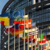 Banderas de la Unión Europea, Ucrania y los Estados miembros ondeando frente al edificio acristalado del Parlamento Europeo, con un cielo despejado al fondo.