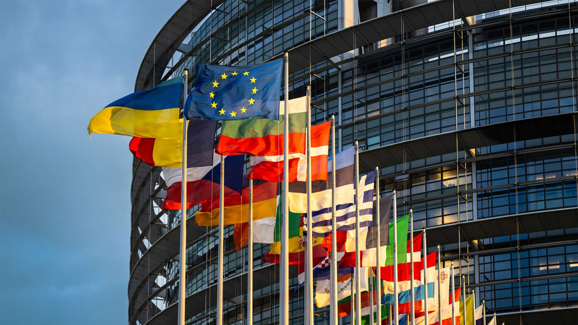 Banderas de la Unión Europea, Ucrania y los Estados miembros ondeando frente al edificio acristalado del Parlamento Europeo, con un cielo despejado al fondo.
