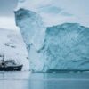 Enorme iceberg de tonos azulados junto a un buque de expedición que navega por las aguas del Ártico en Groenlandia, con un paisaje costero cubierto de nieve al fondo.