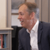 Michael Krake, con traje azul y camisa blanca, habla durante una entrevista sentado de perfil. Al fondo se ve una estantería con libros, una pequeña figura decorativa y una puerta a la derecha.