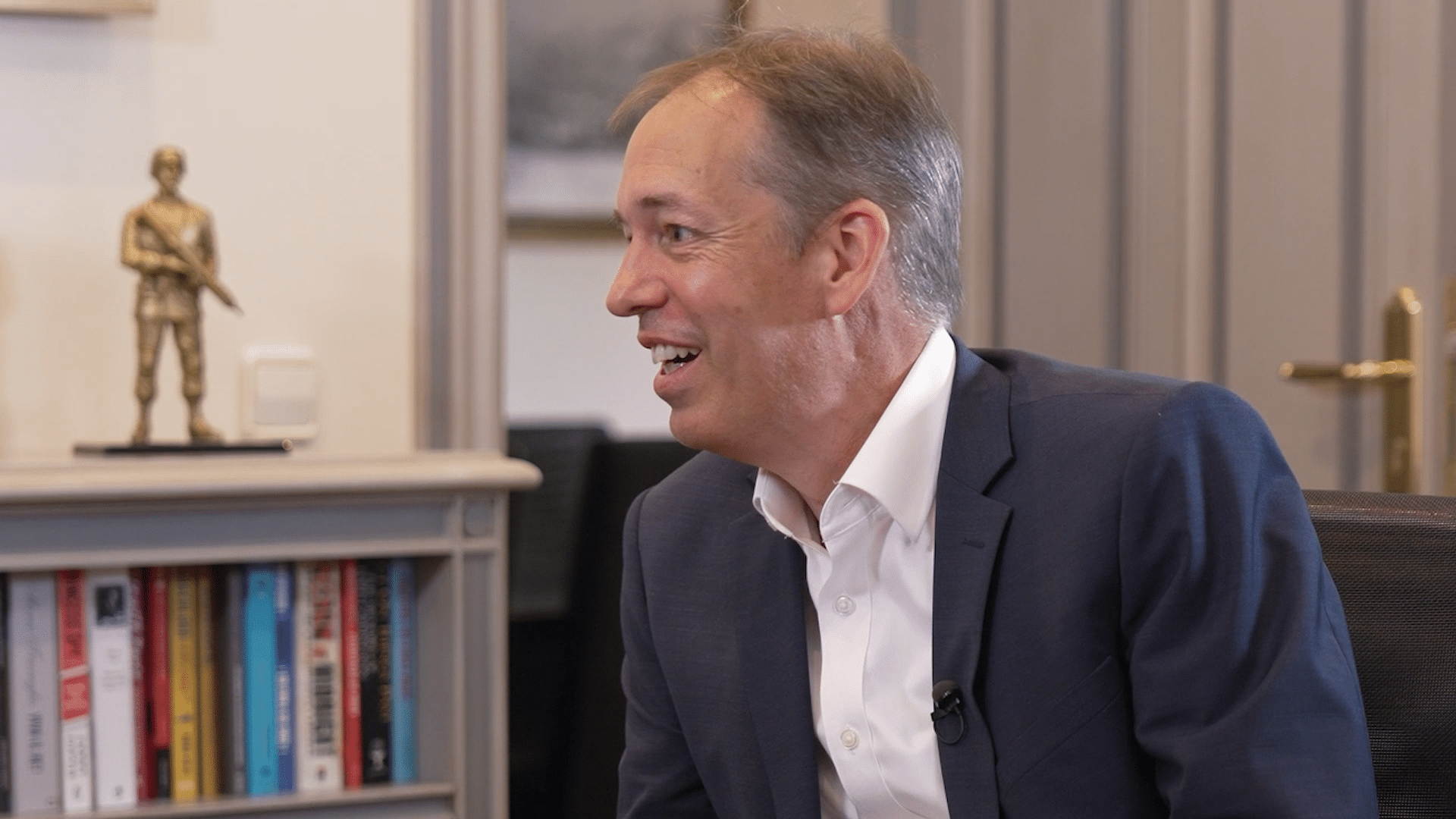 Michael Krake, con traje azul y camisa blanca, habla durante una entrevista sentado de perfil. Al fondo se ve una estantería con libros, una pequeña figura decorativa y una puerta a la derecha.