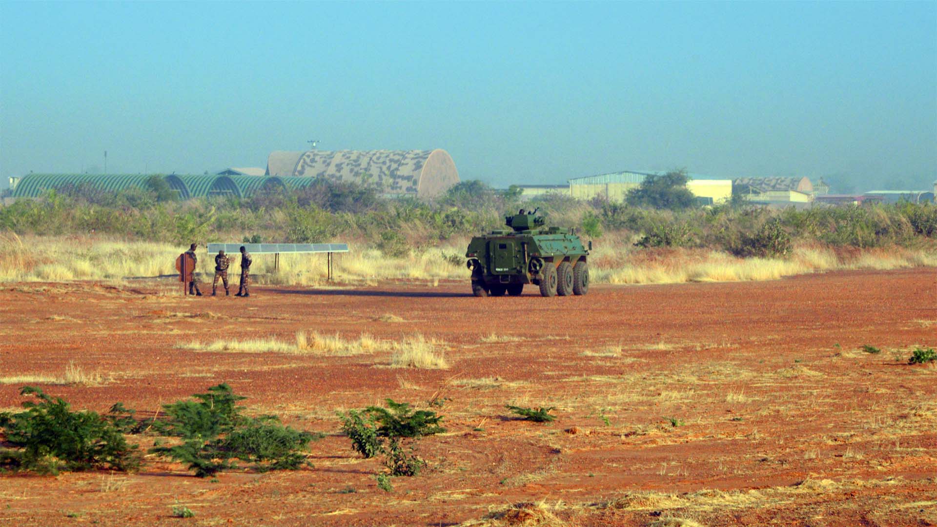 Vehículo blindado y varios soldados en una zona militar del aeropuerto internacional Diori Hamani, en Niamey, Níger, con hangares al fondo y terreno árido en primer plano. El cielo es claro aunque hay un ambiente empolvado.
