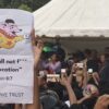 Una multitud de jóvenes congregados con las manos levantadas sosteniendo sus móviles grabando el discurso de una representante del Partido Popular de las Cucarachas (CPJ), quien está en una carpa junto a un grupo de personas. En primer plano, un cartel de Dinkan (un superhéroe en forma de ratón) con la frase “Thou shall not f*** with education” (No te meterás con la educación) y debajo “In Dinkan we trust” (En Dinkan confiamos), el lema la religión paródica llamada dinkoismo.
