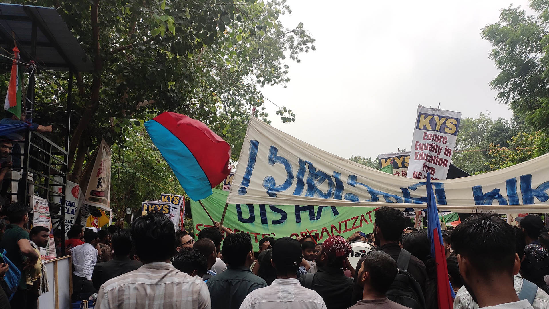 Una multitud de jóvenes estudiantes congregados con pancartas y banderas en una calle arbolada del parque en Jantar Mantar (Nueva Delhi, India) tras la lluvia, durante las protestas por la filtración de las preguntas del examen nacional NEET UG del 20 de julio de 2026.