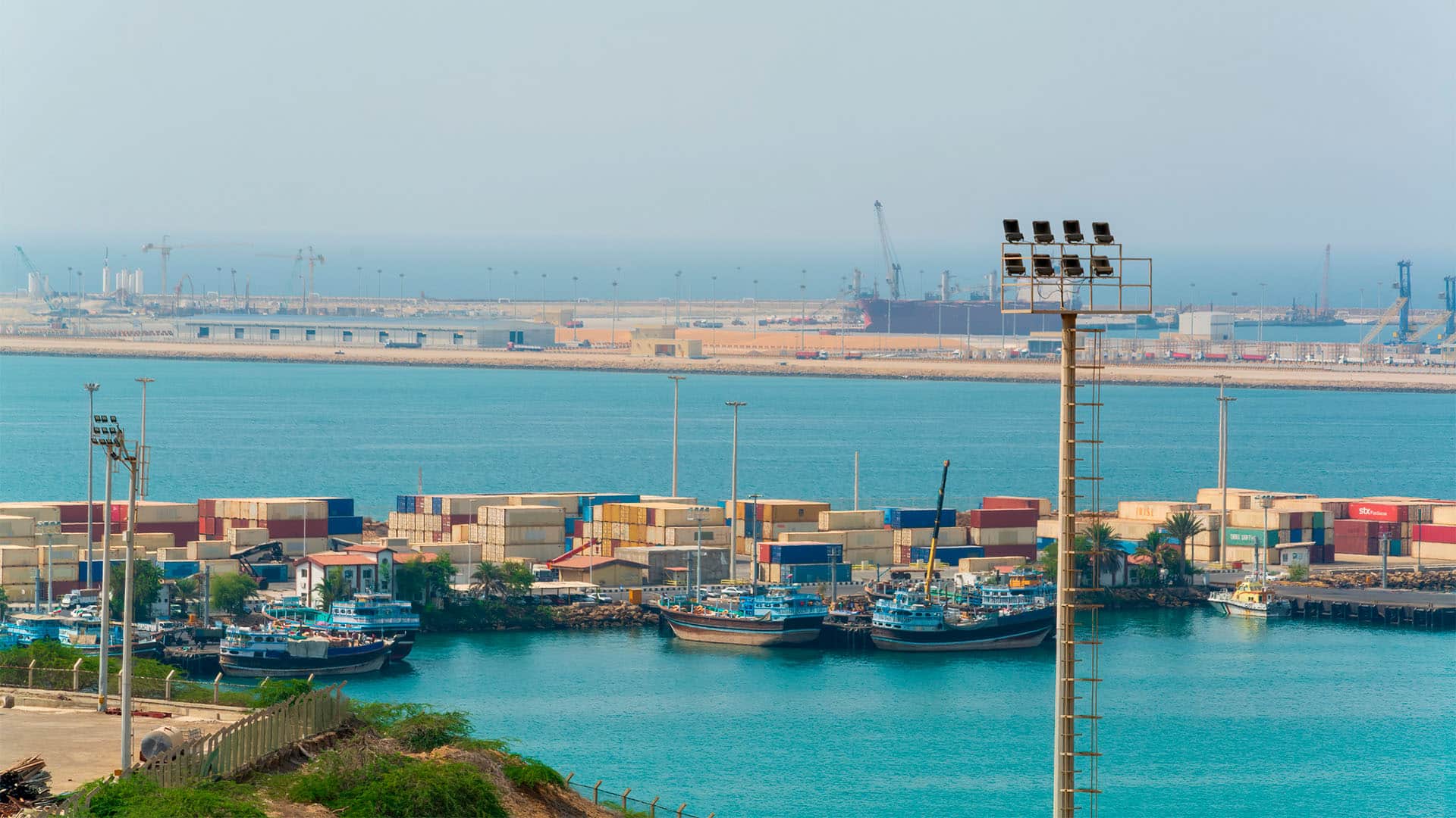 Vista panorámica del puerto de Shahid Beheshti (Chabahar, Irán), con barcos pesqueros, contenedores, grúas e instalaciones portuarias junto al golfo de Omán, bajo un cielo despejado.