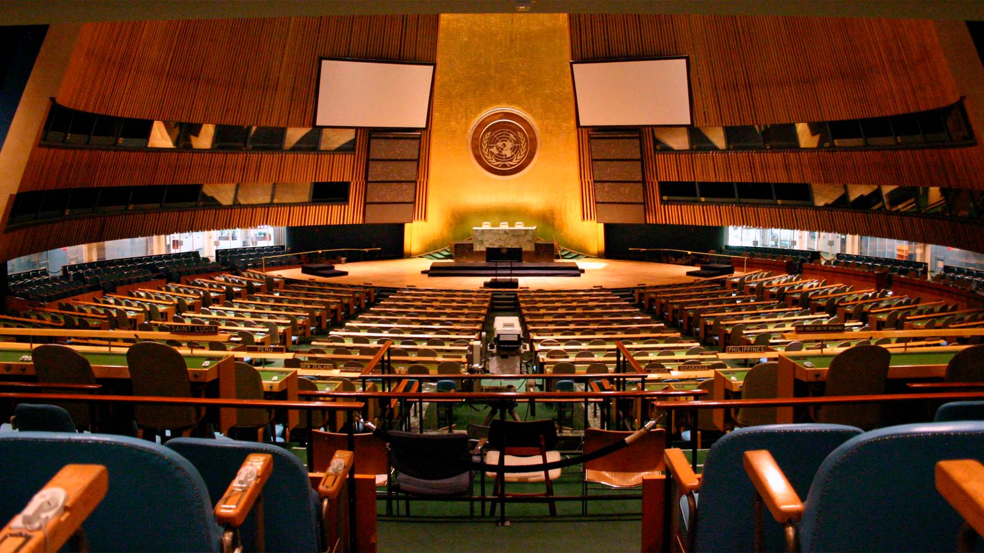 Vista panorámica del salón vacío de la Asamblea General de las Naciones Unidas, con filas de asientos y pupitres de los Estados miembros frente a la tribuna central y el emblema de la ONU.