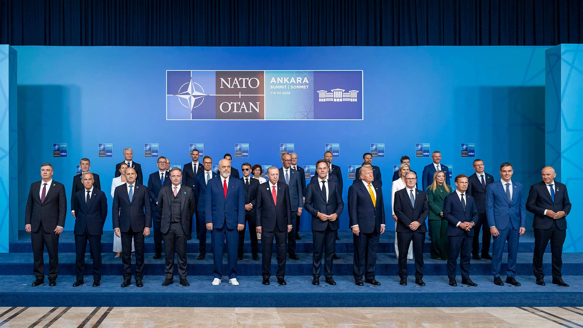 Fotografía de grupo de la cumbre de la OTAN en Ankara. Varias decenas de líderes y representantes posan de pie en tres filas sobre una tarima azul, con un fondo institucional en tonos azules y el cartel “NATO OTAN Ankara Summit 7-8 VII 2026” en la parte superior.