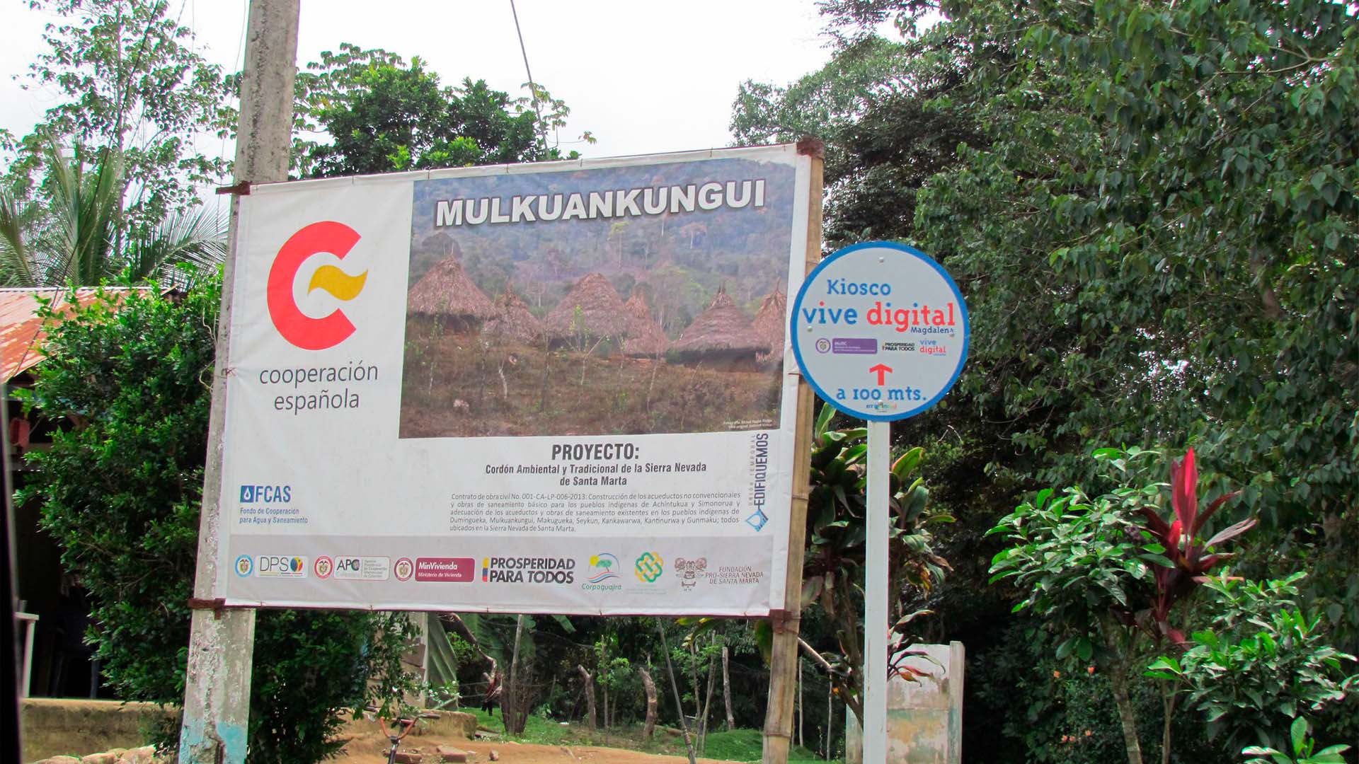 Cartel de la Cooperación Española sobre un proyecto de agua y saneamiento en comunidades de la Sierra Nevada de Santa Marta, Colombia rodeado de vegetación.