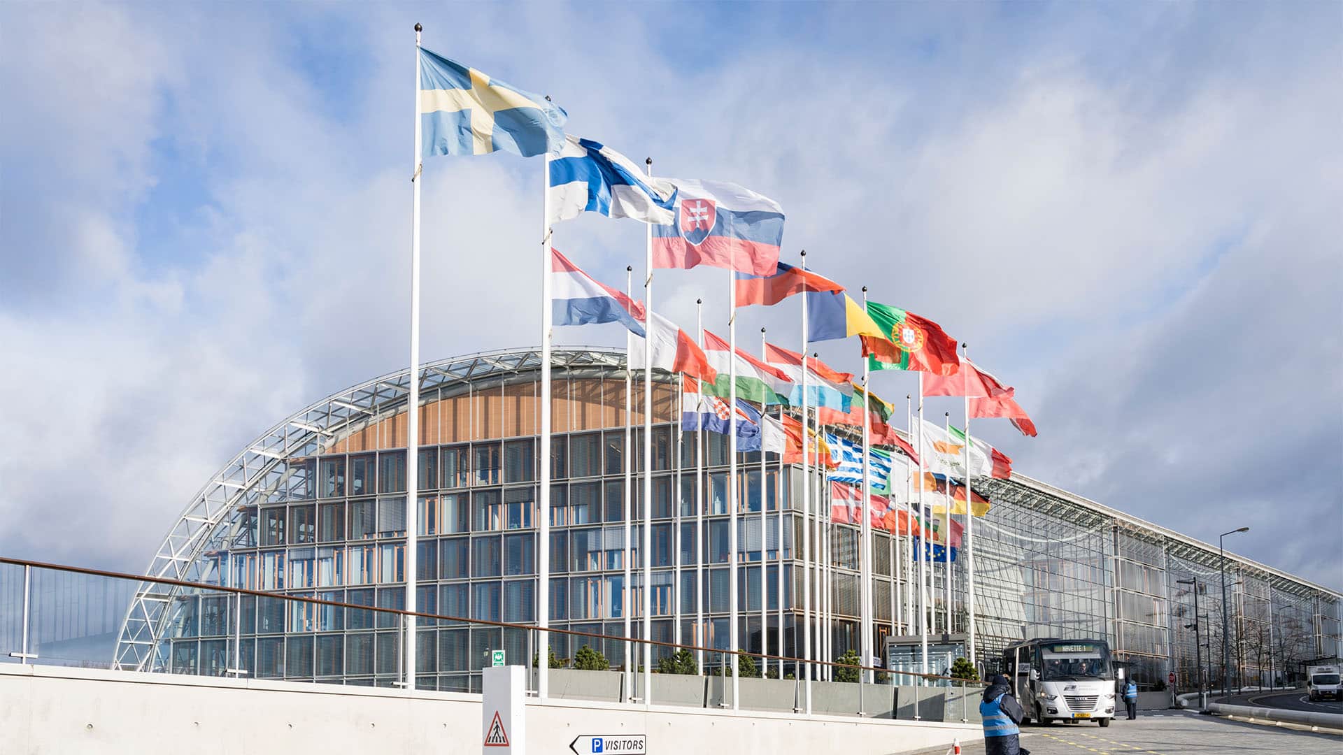 Exterior view of the European Investment Bank Group headquarters in Luxembourg’s Kirchberg district, with a row of European national flags in front of the curved glass-and-steel building.