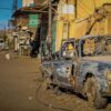 Calle de la ciudad de Omdurmán (Sudán) con edificios de fachada deteriorada y un automóvil calcinado y abandonado en primer plano junto a un muro dañado. Al fondo, otros vehículos, postes eléctricos y comercios bajo luz diurna, en un entorno desgastado y con infraestructuras destruidas.
