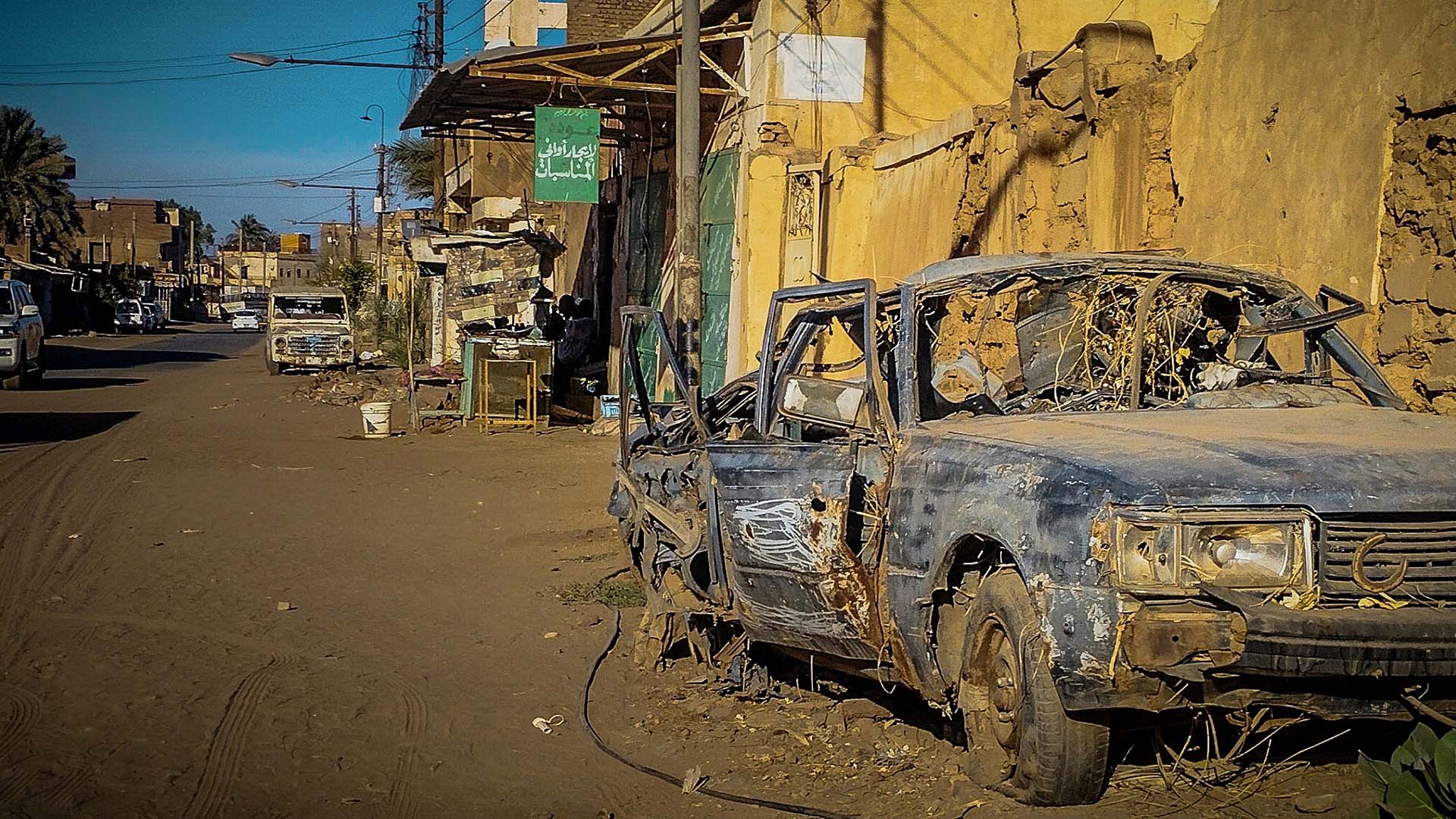 Calle de la ciudad de Omdurmán (Sudán) con edificios de fachada deteriorada y un automóvil calcinado y abandonado en primer plano junto a un muro dañado. Al fondo, otros vehículos, postes eléctricos y comercios bajo luz diurna, en un entorno desgastado y con infraestructuras destruidas.