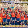 Jugadores de la sección española de fútbol, vestidos con el uniforme rojo y azul, celebran la victoria de la Copa Mundial de la FIFA 2026 sobre el podio del MetLife Stadium repleto de público, mientras cae confeti dorado alrededor. En el centro, el capitán de la sección Rodrigo Hernández Cascante (“Rodri”) levanta el trofeo dorado.